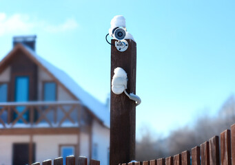 Winter CCTV camera, surveillance, security camera on a snow-covered pole and fence in a country house. Motion sensor burglar alarm in the house