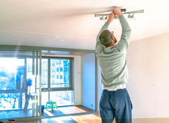 a young man, a newcomer in an empty new apartment against the background of a balcony window flooded with light, prepares lighting and other preparing for moving to a new home