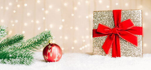 Festive composition with fir tree branch, red ball decor and golden gift box with red velvet ribbon on snow with warm bokeh background. Merry Xmas and Happy new year wide banner. Selective focus.