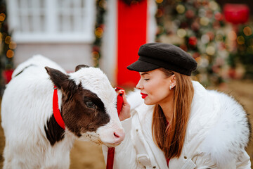 Portrait of young pretty lady in white winter jacket and black cap posing with small bull against Christmas outdoor decor.
