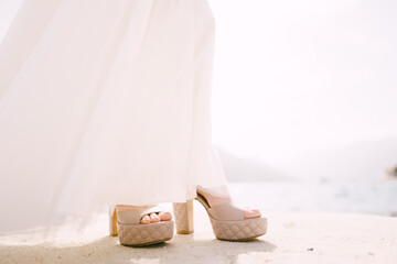 Bride legs in a white dress and beige high-heeled shoes on the pier. Close-up
