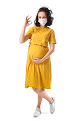 Young pregnant woman in medical mask showing OK on white background