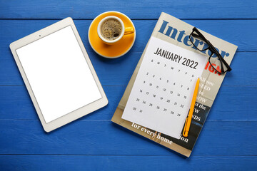 Paper calendar for January 2022, magazine, eyeglasses, tablet computer and cup of coffee on color wooden background