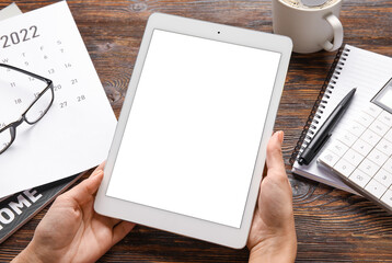 Female hands with tablet computer, paper calendar for 2022 year and stationery supplies on wooden background
