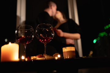 Valentine's day festive dinner, burning candles and wine glasses on the table, couple in love kissing on the background, date in the apartment