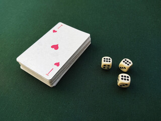 The ace of hearts lies on top of the deck of playing cards. Nearby are three bone dice with the name six on the green material of the game table. The concept of luck and demonstration of gambling, spo