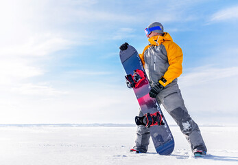 Fototapeta premium Young man holds a snowboard in his hands against the background of a winter landscape. Male in ski goggles and overalls with his board on white snow on a sunny frosty day go in for sports.