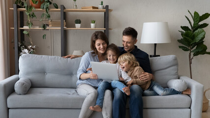 Cheerful millennial parents resting with gen Z kids on couch, making video call, watching movie...