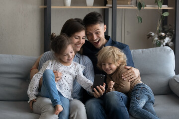 Obraz premium Cheerful daughter girl taking selfie on smartphone with beloved family. Happy couple of parents and two sibling kids relaxing on sofa, posing for self portrait, hugging, smiling, laughing on couch