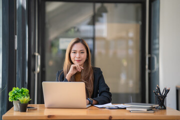 Portrait of a young businesswoman using a laptop, smiling and looking at camera while sitting at office desk in modern office.