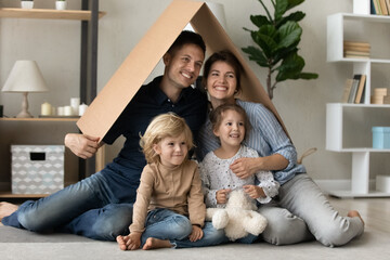 Happy young family couple and two cute sibling kids holding cardboard roof over heads, imitating...