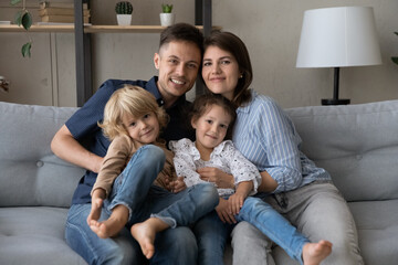 Fototapeta premium Happy millennial couple of parents holding sweet two sibling kids in arms with love, care, resting on couch, relaxing in living room, enjoying family leisure time with cute children. Family portrait