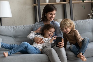 Happy mom and cheerful excited two kids making video call on smartphone, taking funny selfie photo, watching online media on mobile phone, holding cellphone, relaxing on comfortable couch at home