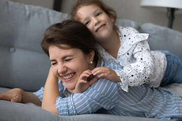 Cheerful laughing mother and kid resting on couch at home, playing, having fun. Happy mom lying on sofa, piggybacking sweet preschool girl, enjoying active games with child. Motherhood, family time