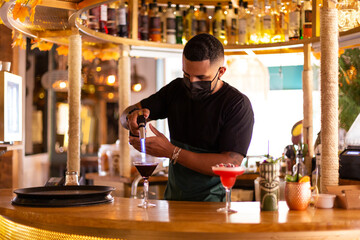 Latin American bartender with face mask working behind the bar in a modern cocktail bar. Space for...