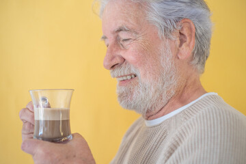 Close up on attractive bearded senior man holding a glass of coffee and milk, cappuccino, standing on yellow background
