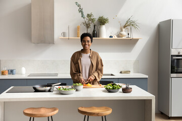 Portrait of smiling young African American woman in eyewear preparing healthy food in modern kitchen, chopping fresh organic vegetables for salad looking at camera, culinary activity concept.