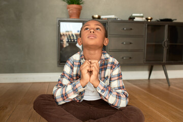 Cute African American boy of 8 years old in flannel shirt and velvet jeans looking up pressing fists, praying for his mom return home from work ASAP, sitting on floor alone in apartment