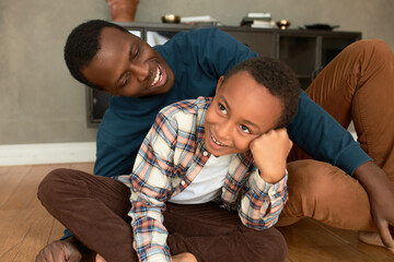 African father cuddling his 9-years-old son while fooling around on floor in living-room against...