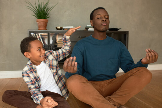 Smiling cheerful naughty brown-skinned casually dressed boy of 8-years-old disturbing his dad meditating in lotus posture, opening one eye, to catch little prankster. Human relationships - Powered by Adobe