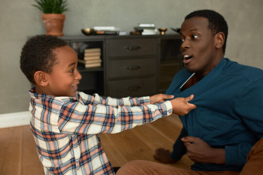 Father and son relationships. Indoor picture of kid and dad fighting and struggling as joke, boy pulling his jumper, both smiling, having joyful face expressions, looking at each other