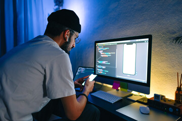 Young man mobile developer writes program code on a computer and check app in smartphone,...