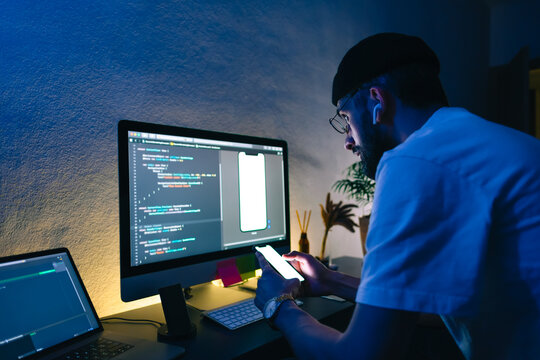 Young man mobile developer writes program code on a computer and check app in smartphone, programmer work in home office.