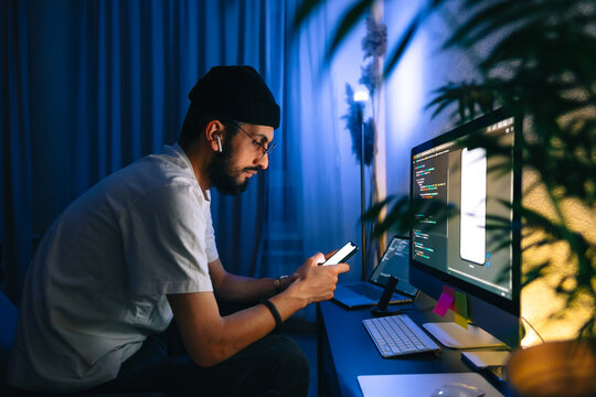 Young man mobile developer writes program code on a computer and check app in smartphone, programmer work in home office.