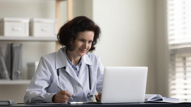 Smiling Positive Mature Physician Doctor Meeting Patient Virtually, Giving Online Remote Video Consultation On Laptop, Writing Medical Records, Listening Complaints. Healthcare Concept