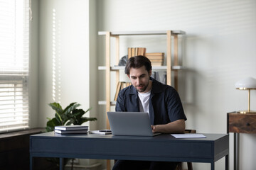 Focused millennial entrepreneur working at laptop at office table. Young business man in casual, employee, worker using computer at workplace, typing, chatting online, making video call