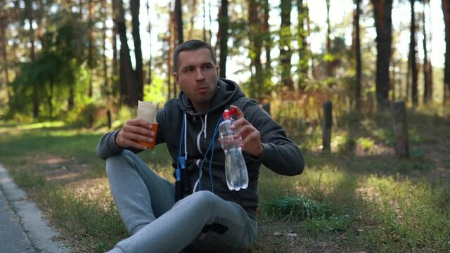 A man in a park or forest on a roadside sits and eats a roll and drinks water from a plastic bottle after a hard street workout