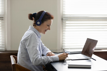 Happy elder student in headphones watching learning webinar on laptop, virtual workshop, writing...