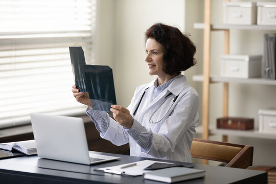 Smiling busy mature female doctor studying xray radiography images. General practitioner, surgeon woman satisfied with good screening test result reviewing scan of patient bones. Healthcare concept
