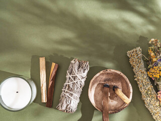 Items for spiritual cleansing - sage and various herbs bundles, palo santo incense sticks and candle on green background with shadows.