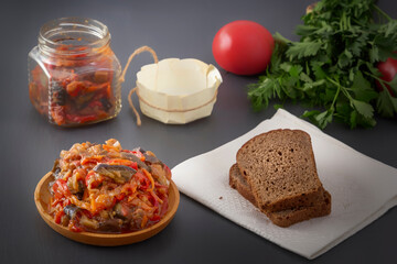 Stewed eggplant with vegetables in a wooden plate and a piece of bread on a white napkin on a gray table against the background of a glass jar, tomato and greens. Homemade.
