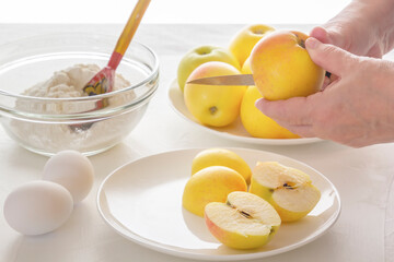 Female hands cut apples for a pie on the background of a cup with flour and chicken eggs on a white table