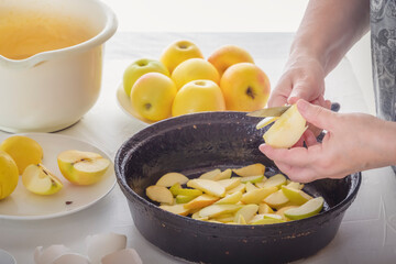 Female hands cut apples for a pie in an old cast-iron pan on the background of a cup with dough and broken chicken eggs on a white kitchen table
