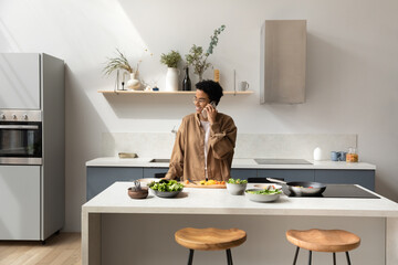 Happy young attractive African American woman in glasses holding pleasant cellphone call conversation, communicating with friends inviting for domestic party, distracted from preparing food in kitchen