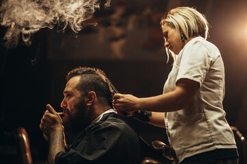 Young bearded man with a mohawk getting haircut by hairdresser and smoking a cigarette
