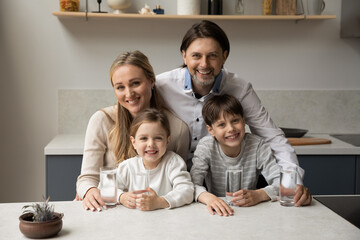Obraz na płótnie Canvas Portrait of smiling bonding young couple parents and little children siblings holding glasses of fresh pure mineral water in hands, refreshing in modern kitchen, daily healthcare family habit concept.
