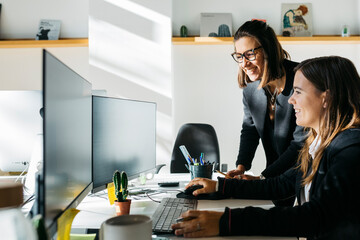 Smiling businesswoman listening to a collegue, in front of a computer, at desk, in office. They are in smart casuals.