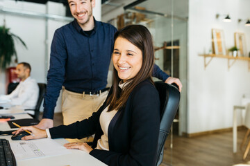 Smiling businesswoman listening to a collegue, in front of a computer, at desk, in office. They are in smart casuals.