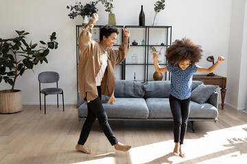 Full length overjoyed millennial African American mother or babysitter dancing to favorite energetic disco music with happy small adorable child barefoot on warm wooden floor in modern living room.