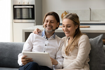 Happy laughing affectionate bonding young family couple using digital computer tablet applications, watching funny photo video content in social network, shopping online, tech addiction concept.