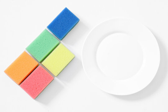 Five Multi-colored Dishwashing Sponges And A Clean White Round Plate On A White Table. View From Above. The Concept Of Efficient Dishwashing And Cleaning In The Kitchen