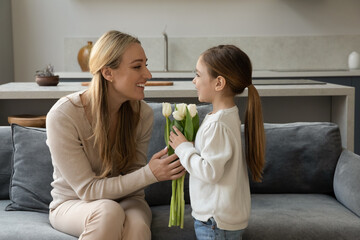 Happy young mother feeling excited from getting flowers from little cute child daughter. Cheerful small kid girl presenting gift bouquet to smiling millennial mum, congratulating with birthday at home