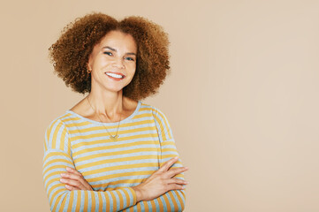Studio portrait of beautiful middle age woman isolated on beige background, wearing grey and yellow stripe t-shirt, mixed race lady with curly hair - 473126282