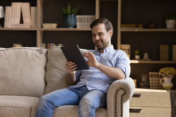 Happy young man holding digital device, resting on couch at home, using online virtual app on tablet, browsing internet, chatting, reading book, watching multimedia content, laughing