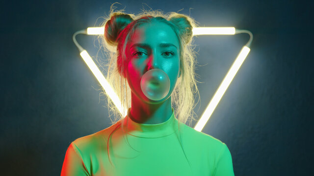 modern young girl blowing gum bubble in changing colorful light on background with triangular neon lamp
