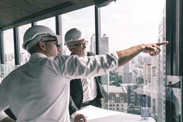 Two engineering working together in their new project. They are wearing suit , tie and white hard hat. They are looking at their design on the paper on the wooden table